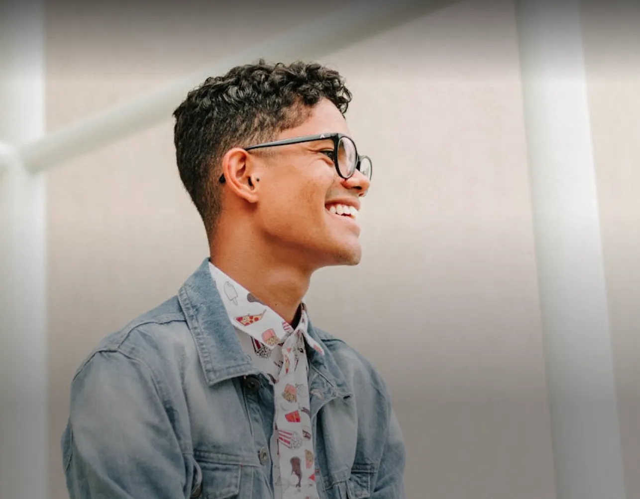 Young man with curly hair and glasses smiling and looking to the side wearing a denim jacket and printed shirt.